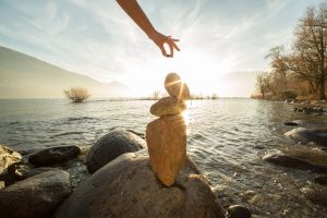 A hand carefully balancing stones beside a lake at sunrise